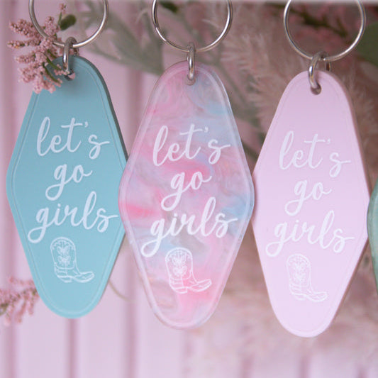 Three keychains with 'let's go girls' text on a pink background