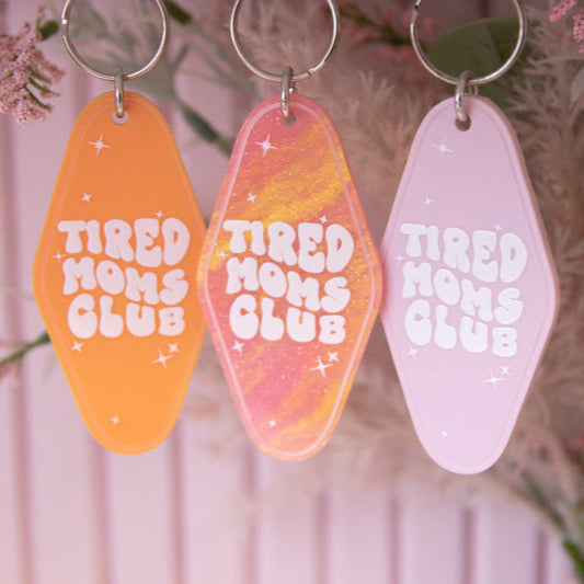 Three 'Tired Moms Club' keychains in orange, orange and pink marble, and pink on a floral background.